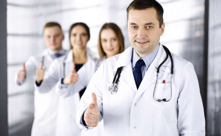 Group of professional doctors are standing as a team with thumbs up in a hospital office, ready to help their patients. Medical help, insurance in health care and medicine conceptの写真素材