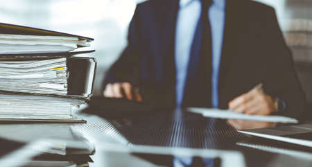 Binders with papers are waiting to be processed by man accountant in blue blazer staying at home during pandemic. Taxes and audit concept. Internal Revenue Service inspector checking financial document.の写真素材