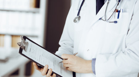Unknown male doctor standing and working with clipboard of medication history records in clinic at his working place, closeup. Perfect medical service in hospital. Medicine and healthcare conceptの写真素材
