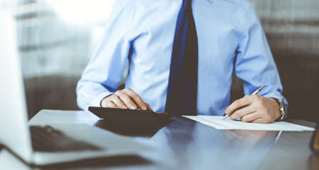 Unknown man accountant in blue shirt use calculator and computer with holding pen on while staying...
