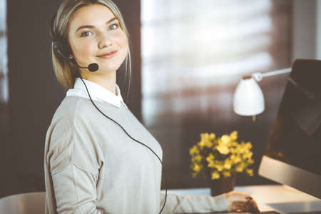 Diverse people group in sunny call center. Blonde business woman talking by headset while sitting in modern officeの写真素材