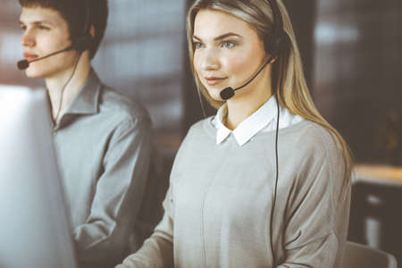Diverse people group in sunny call center. Blonde business woman talking by headset while sitting in modern officeの写真素材