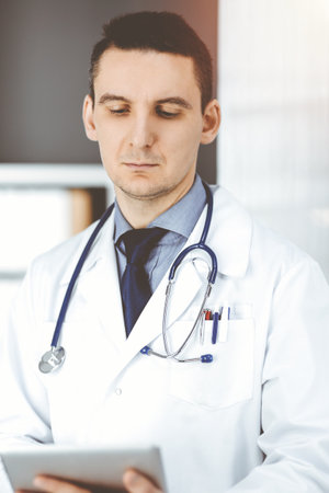 Friendly male doctor using tablet computer in clinic near his working place. Perfect medical service in hospital. Medicine and healthcare conceptの写真素材