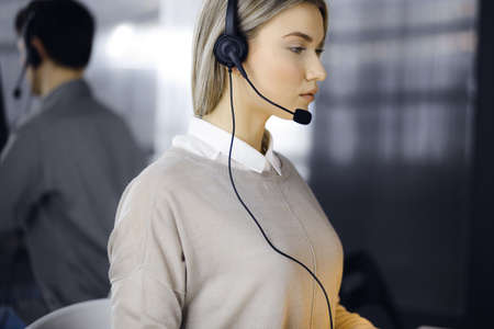 Blonde business woman talking by headset while sitting in modern call center office. Telemarketing and customer service conceptの写真素材