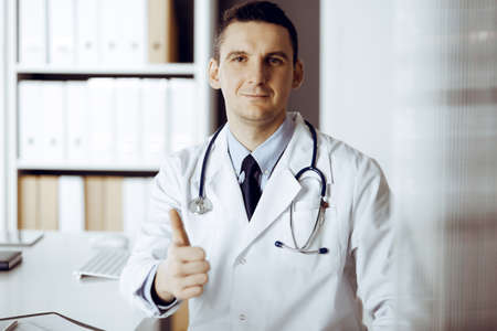 Friendly male doctor sitting and showing OK sign at his working place in clinic. Perfect medical service in hospital. Medicine and healthcare conceptの写真素材