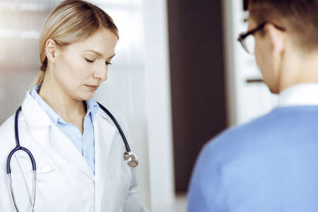 Friendly female doctor and patient man discussing current health examination while sitting in sunny clinic. Perfect medical service in hospitalの写真素材