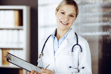 Friendly female doctor standing in sunny clinic. Portrait of cheerful smiling physician. Perfect medical service in hospitalの写真素材