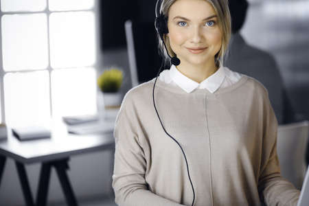 Blonde business woman talking by headset while sitting in modern call center office. Telemarketing and customer service conceptの写真素材