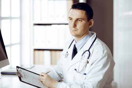 Friendly male doctor sitting and working with clipboard of medication history records in sunny clinic. Medicine and healthcare conceptの写真素材
