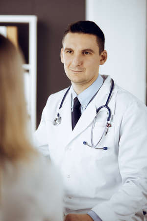 Friendly male doctor and patient woman discussing current health examination while sitting in sunny clinic. Perfect medical service in hospitalの写真素材