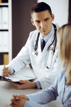 Friendly male doctor and patient woman discussing current health examination while sitting in sunny clinic. Perfect medical service in hospitalの写真素材