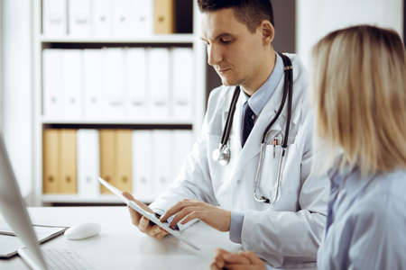 Friendly male doctor and patient woman discussing current health examination while sitting in sunny clinic. Perfect medical service in hospitalの写真素材