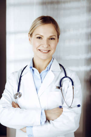Friendly female doctor standing in sunny clinic. Portrait of cheerful smiling physician. Perfect medical service in hospitalの写真素材