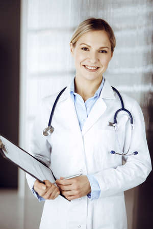 Friendly female doctor standing in sunny clinic. Portrait of cheerful smiling physician. Perfect medical service in hospitalの写真素材