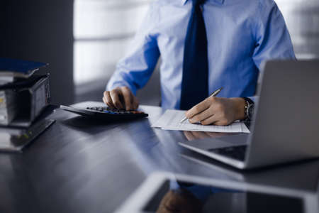 Unknown man accountant in blue shirt use calculator and computer with holding pen on while staying at home during covid pandemic. Taxes and audit conceptの写真素材