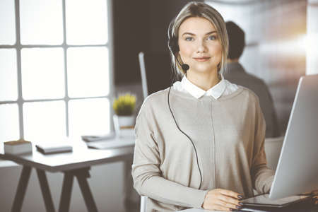 Diverse people group in sunny call center. Blonde business woman talking by headset while sitting in modern officeの写真素材
