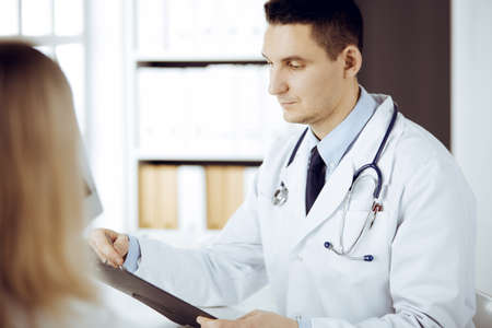 Friendly male doctor and patient woman discussing current health examination while sitting in sunny clinic. Perfect medical service in hospitalの写真素材