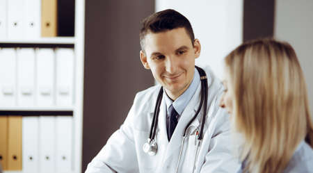 Friendly male doctor and patient woman discussing current health examination while sitting in sunny clinic. Perfect medical service in hospitalの写真素材