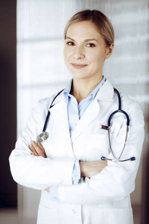Friendly female doctor standing in sunny clinic. Portrait of cheerful smiling physician. Perfect medical service in hospitalの写真素材