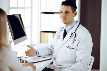 Friendly male doctor and patient woman discussing current health examination while sitting in sunny clinic. Perfect medical service in hospitalの写真素材