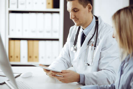 Friendly male doctor and patient woman discussing current health examination while sitting in sunny clinic. Perfect medical service in hospitalの写真素材