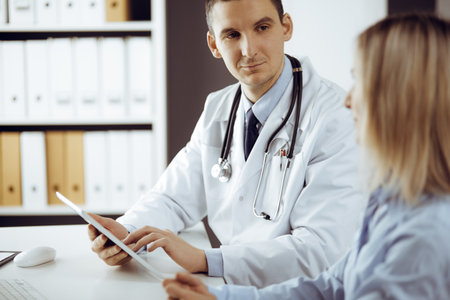 Friendly male doctor and patient woman discussing current health examination while sitting in sunny clinic. Perfect medical service in hospitalの写真素材