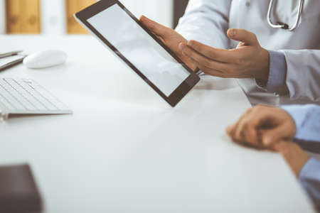 Unknown male doctor and patient woman discussing current health examination while sitting in clinic and using tablet computer, closeup of hands. Medicine and healthcare conceptの写真素材