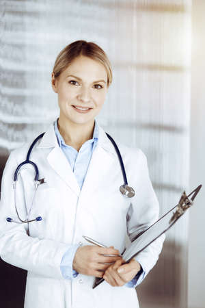 Friendly female doctor standing in sunny clinic. Portrait of cheerful smiling physician. Perfect medical service in hospitalの写真素材