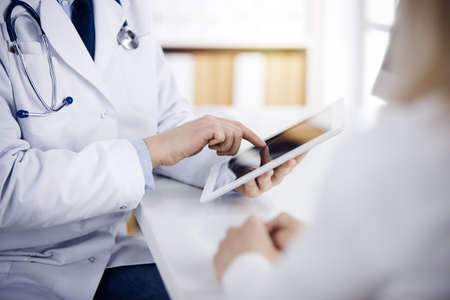 Unknown male doctor and patient woman discussing current health examination while sitting in sunny clinic and using tablet computer. Perfect medical service in hospitalの写真素材