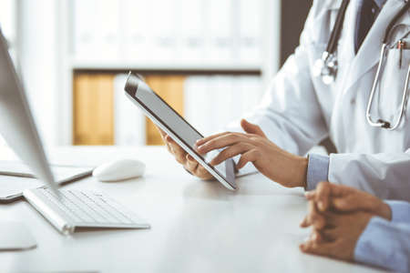 Unknown male doctor and patient woman discussing current health examination while sitting in clinic and using tablet computer, closeup of hands. Medicine and healthcare conceptの写真素材