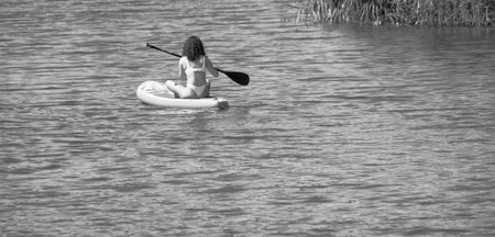 Three people enjoy an active summer day, paddleboarding on inflatable SUP boards along a calm river. In the background, a sandy beach with sunbathers and lush riverside greenery sets a peaceful summer scene. Bright orange boards contrast with the blue ripples of the water, creating a vibrant and carefree atmosphere. Perfect for illustrating concepts of outdoor activities, water sports, travel, and summer vacations. on the water people paddleboarding on the riverの写真素材