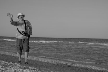 Middle-aged person standing along the seashore in summer with camera and taking pictures of the beauty of nature. In the distance, you can see people, sea, and clouds.の写真素材