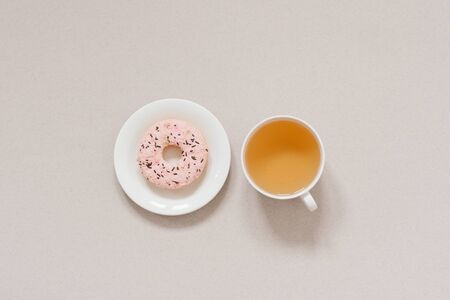 The minimalist banner coffee break. Green tea in a white porcelain Cup and pink donut on a white plate on a beige background. Morning dessertの写真素材