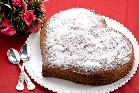 Heart shaped chocolate cake on red tablecloth with two crossed silver little spoons and bouquetの写真素材
