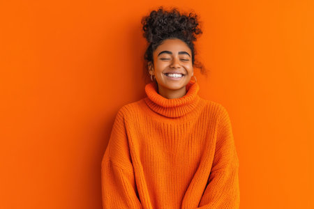 A cheerful young woman wearing an orange sweater stands against a vibrant orange wall, showcasing a joyful smile. The image captures a sense of warmth and positivity, perfect for conveying happiness and style.ai generated.の素材
