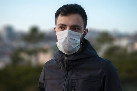 Isolated young man wearing mask to protect himself from the corona virus looks towards with blurred buildings in backgroundの写真素材