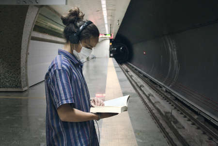 Isolated masked long haired young adult listening to music from his headphone and reading literature book while waiting for the subway to comeの写真素材