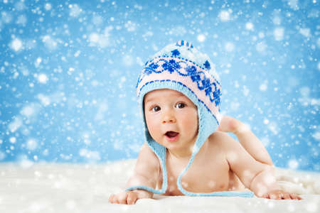 Eight month old baby in knitted winter hat lying on soft blanket with blue background and falling snowの写真素材
