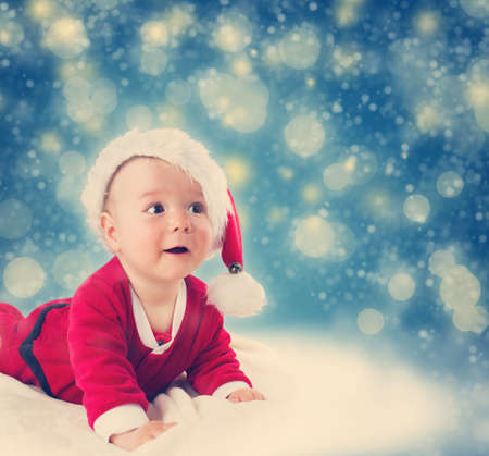 Eight month old baby in santa costume lying on soft blanket with blue background and falling snowの写真素材