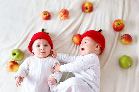 two cute six month old babies lying in hats on soft blanket with apples