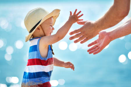 one year old boy playing with father at the beach in straw hat. Child on family vacations at sea. Mans hands and a babyの写真素材