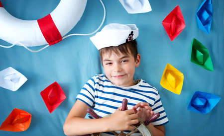 seven years old boy playing in sailor hat. Child with a steering wheel and paper ships on blue...