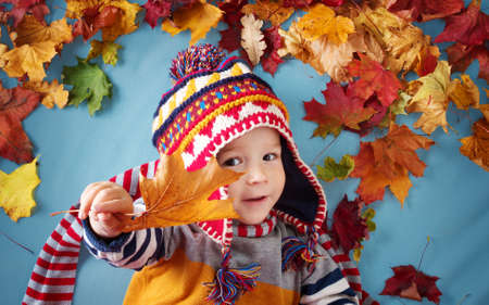 two years old boy dreaming in autumn. Child in hat and scarf and maple leaves on blue backgroundの写真素材