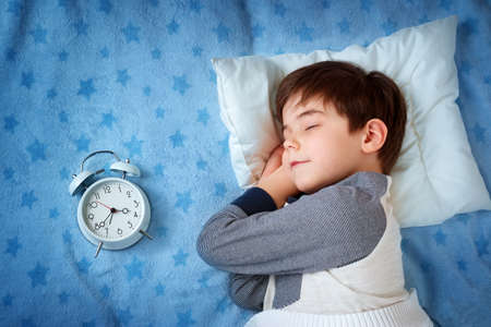 six years old child sleeping in bed on pillow with alarm clock and a teddy bearの写真素材