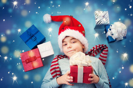 Happy child on blue blanket in red hat holding gift box. Cute boy in christmas cap with many presentsの写真素材