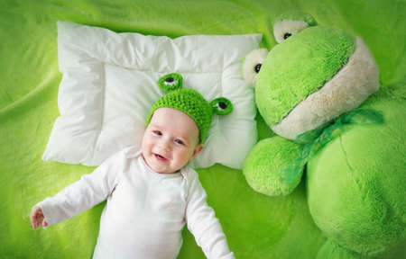 little boy in knitted animal hat lying on soft green blanket with frog softtoy. Cute 5 month old baby on the pillow in bedの写真素材