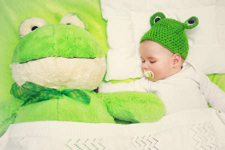 little boy in knitted animal hat sleeping on soft green blanket with frog soft toy. Cute 5 month old baby on the pillow in bedの写真素材