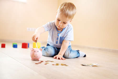 two years old child sitting on the floor and putting money into a piggybankの写真素材