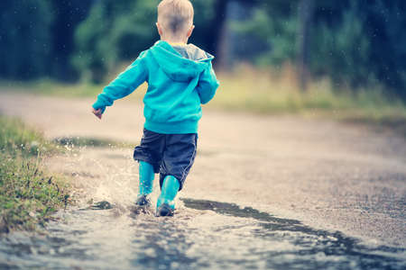 Child walking in wellies and jumping in puddle on rainy weather. Boy under rain in summer outdoorsの写真素材