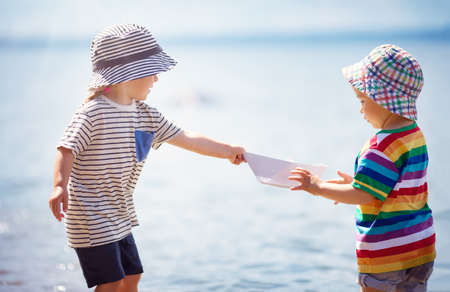 Girl and boy playing on the beach in summer hats and holding paper shipsの写真素材